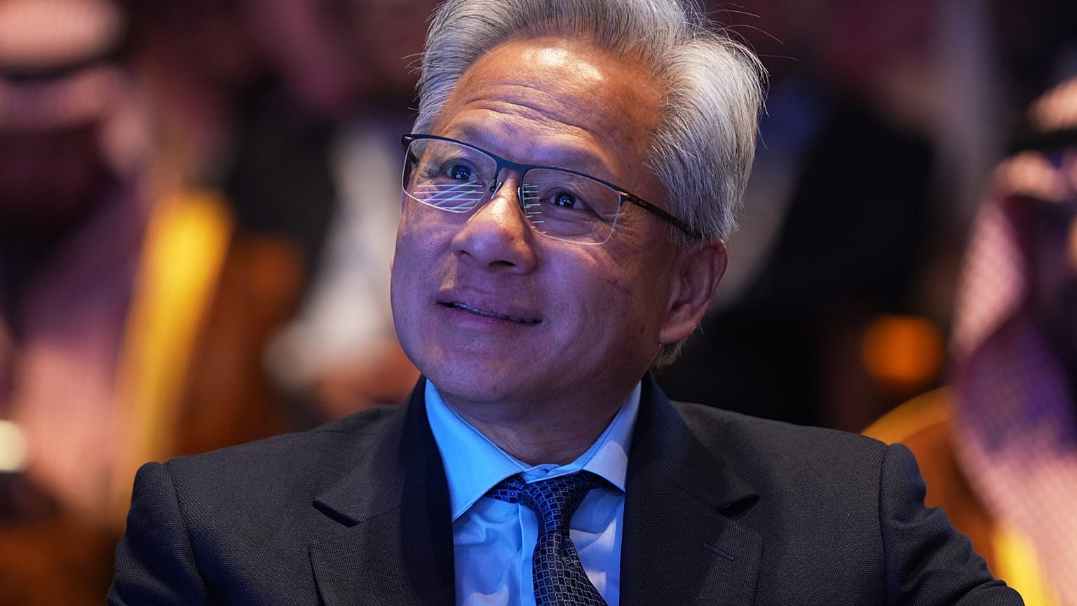 FILE. Nvidia CEO Jensen Huang listens as President Donald Trump speaks during the Saudi Investment Forum at the Kennedy Center. 19 Nov. 2025.